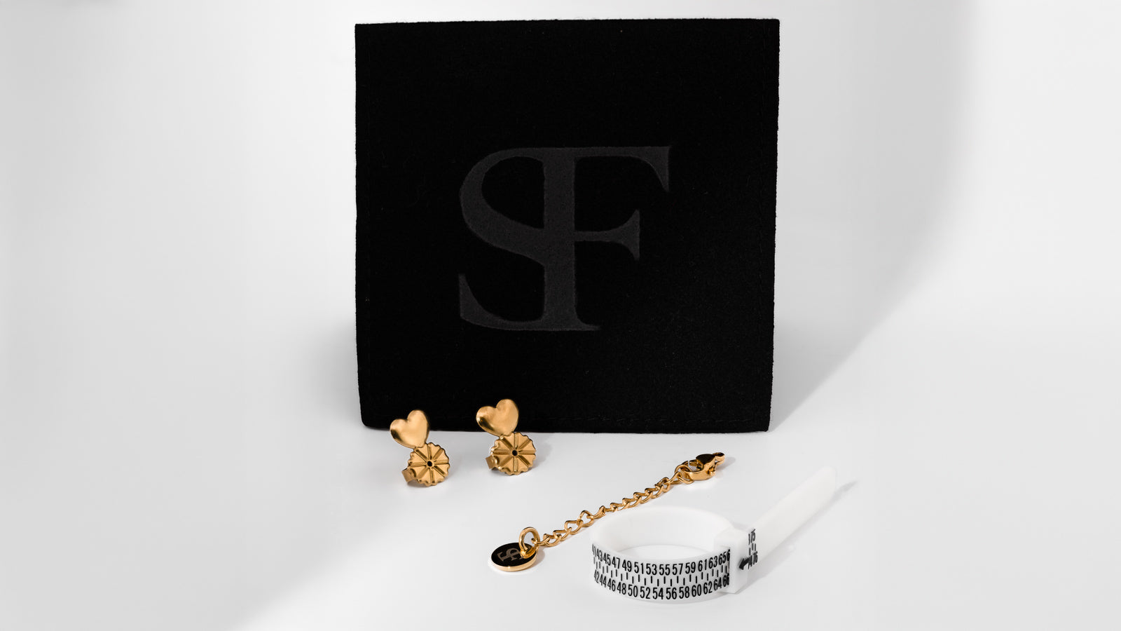 Elegant svart ask med SF-logotyp från Susanna Falken Jewellery, tillsammans med en smyckespåse, hudvänliga guldfärgade örhängen med Stadig Snurrebuss, en förlängningskedja och ett måttband för fingrar – fotograferat på vit bakgrund.